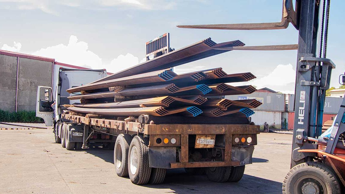 Forklift loading Z-type steel sheet piles onto a flatbed truck at a construction supply yard in Davao, Philippines, with shipping containers visible in the background