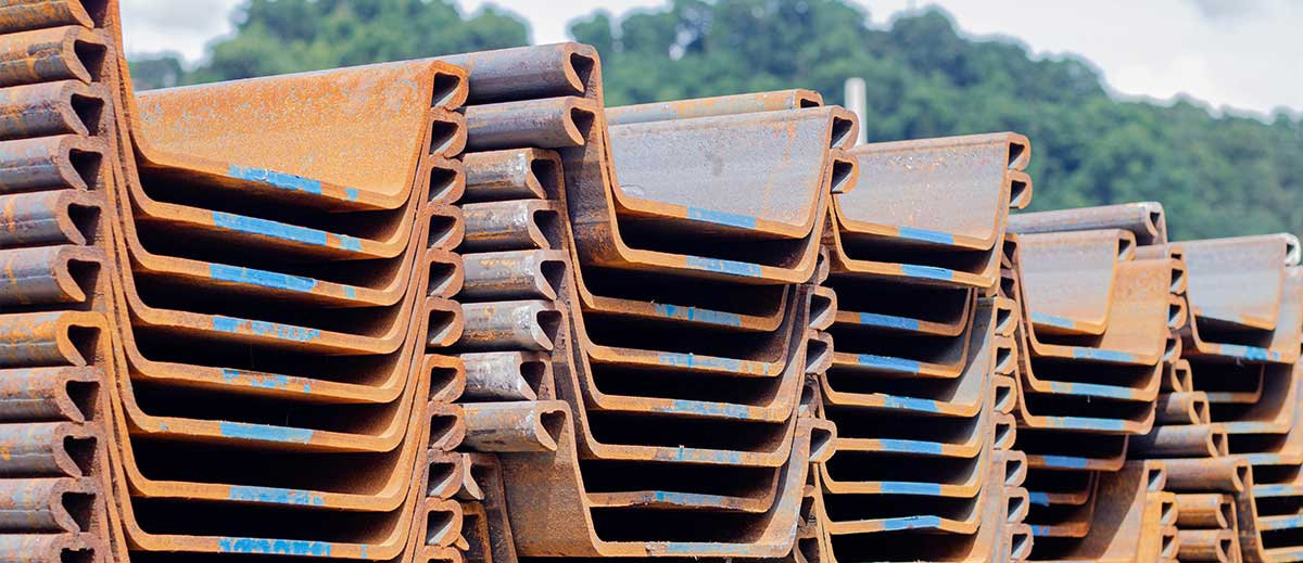 Neatly stacked rows of U-type sheet piles showing rusty orange surfaces with blue markings along the edges, displaying their distinctive U-shaped profiles with interlocking edges against a background of green trees.