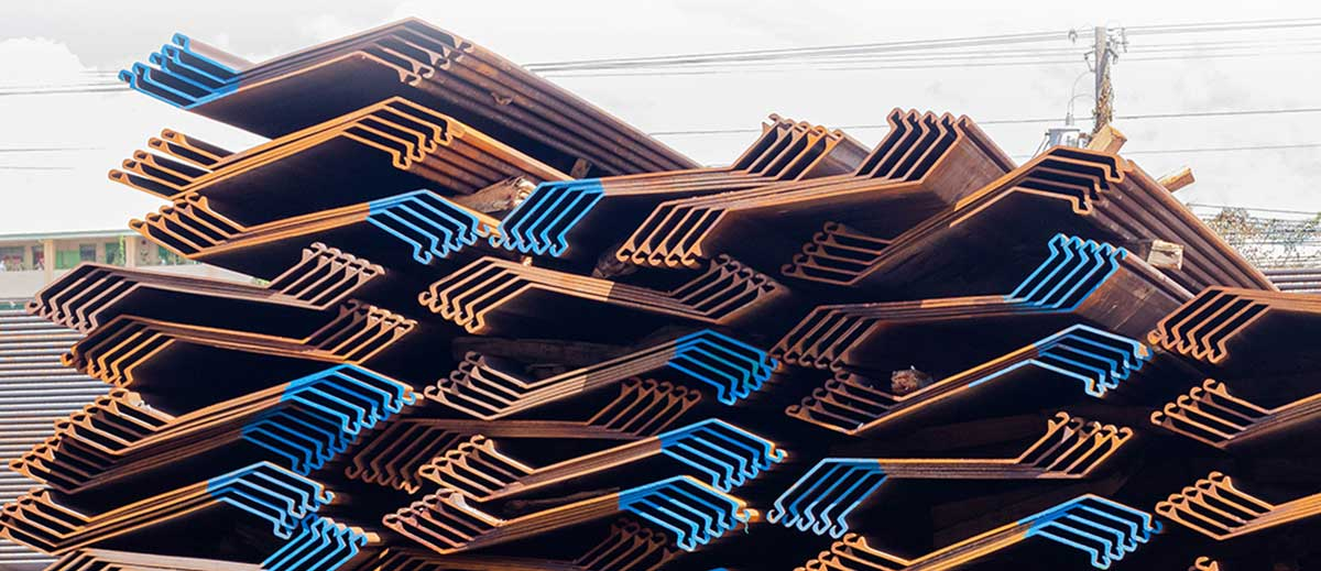A large stack of Z-type sheet piles at a construction materials storage area. The steel sheet piles feature the distinctive Z-shaped profile with interlocking edges. Most are in a rusty brown/orange color with some sections highlighted in blue paint along the edges. The sheet piles are stacked in multiple layers with their interlocking connections visible. Power lines can be seen in the background against a cloudy sky.